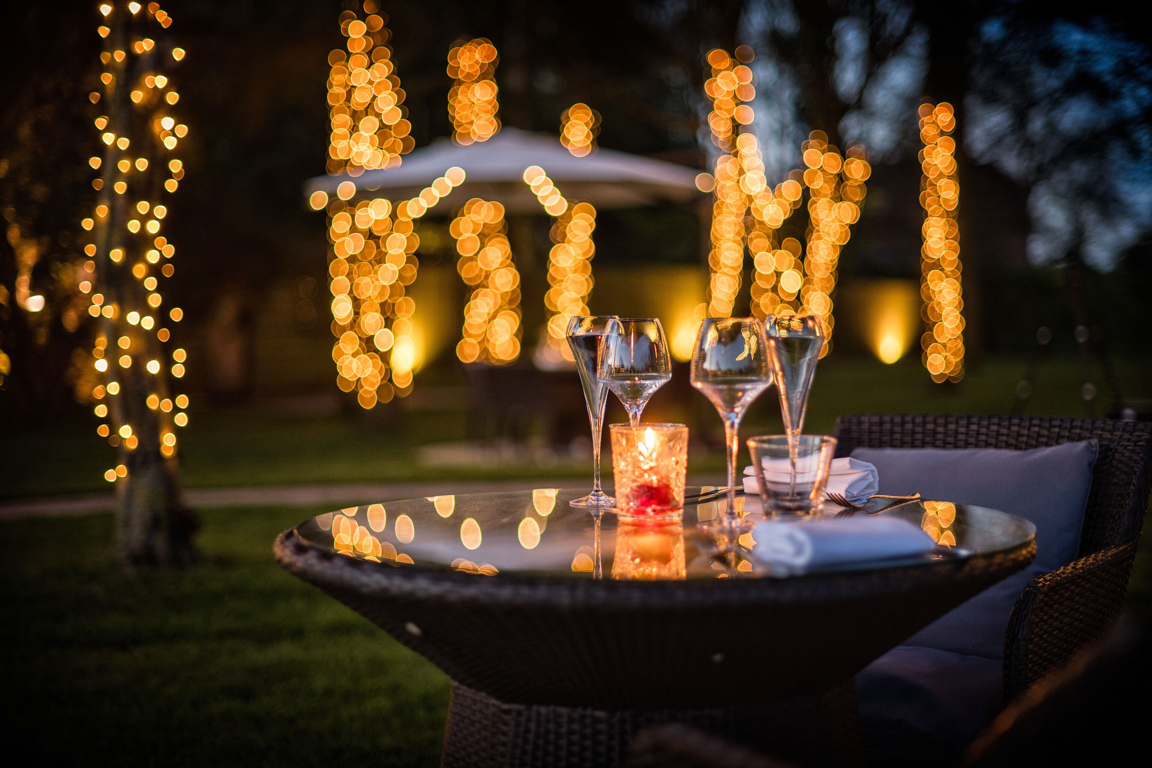 A table with a glass candle, two wine glasses, and a napkin, set outdoors with string lights in the background.