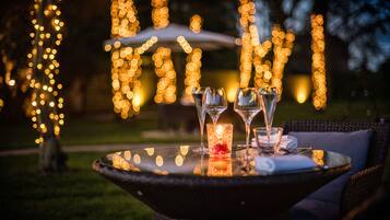 A table with a glass candle, two wine glasses, and a napkin, set outdoors with string lights in the background.
