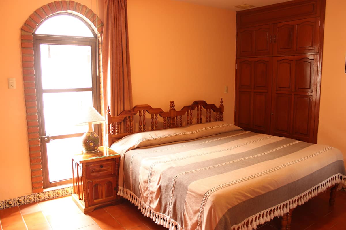 Ein Schlafzimmer mit einem großen Bett, einem Nachttisch aus Holz mit einer Lampe, einem Rundbogenfenster aus Ziegeln und einem Holzschrank.