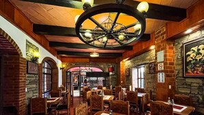 A restaurant interior with brick walls, wooden beams, and a large circular chandelier. Tables are set with chairs and have menus on them.