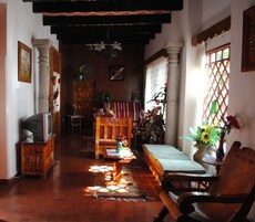 Una sala de estar tradicional con un banco de madera, un sofá y un televisor sobre un mueble. Se ve una ventana con un diseño de celosía y un florero con flores sobre una mesa.