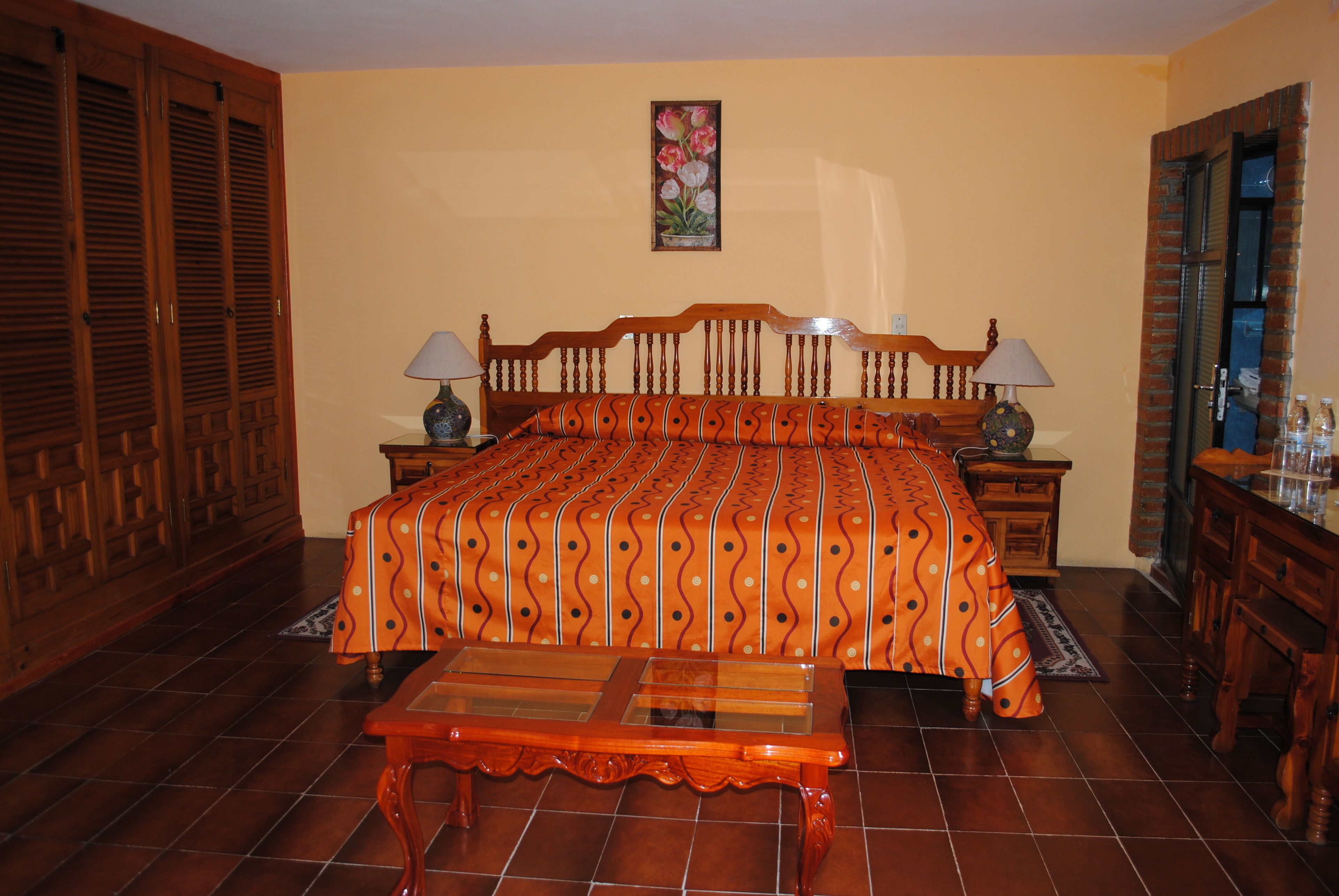 Un dormitorio con una cama de madera, dos mesitas de noche con lámparas, un banco de madera y suelo de baldosas.