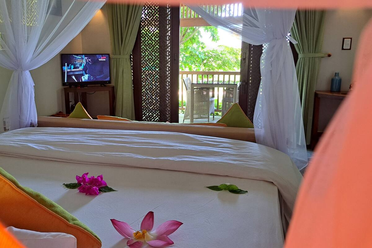 A hotel room with a bed, a television, and a view of greenery outside.