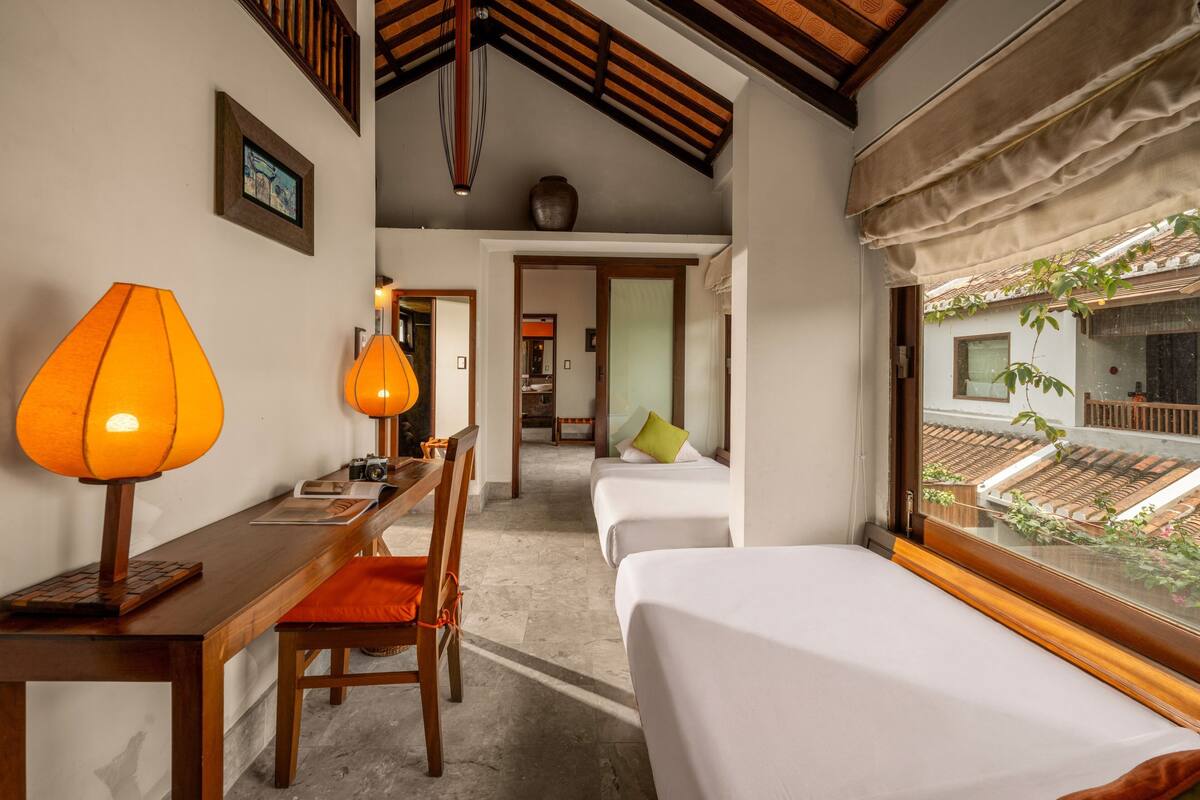 A hotel room with a wooden desk, two beds, a chair, a lamp, and a view of the outside.