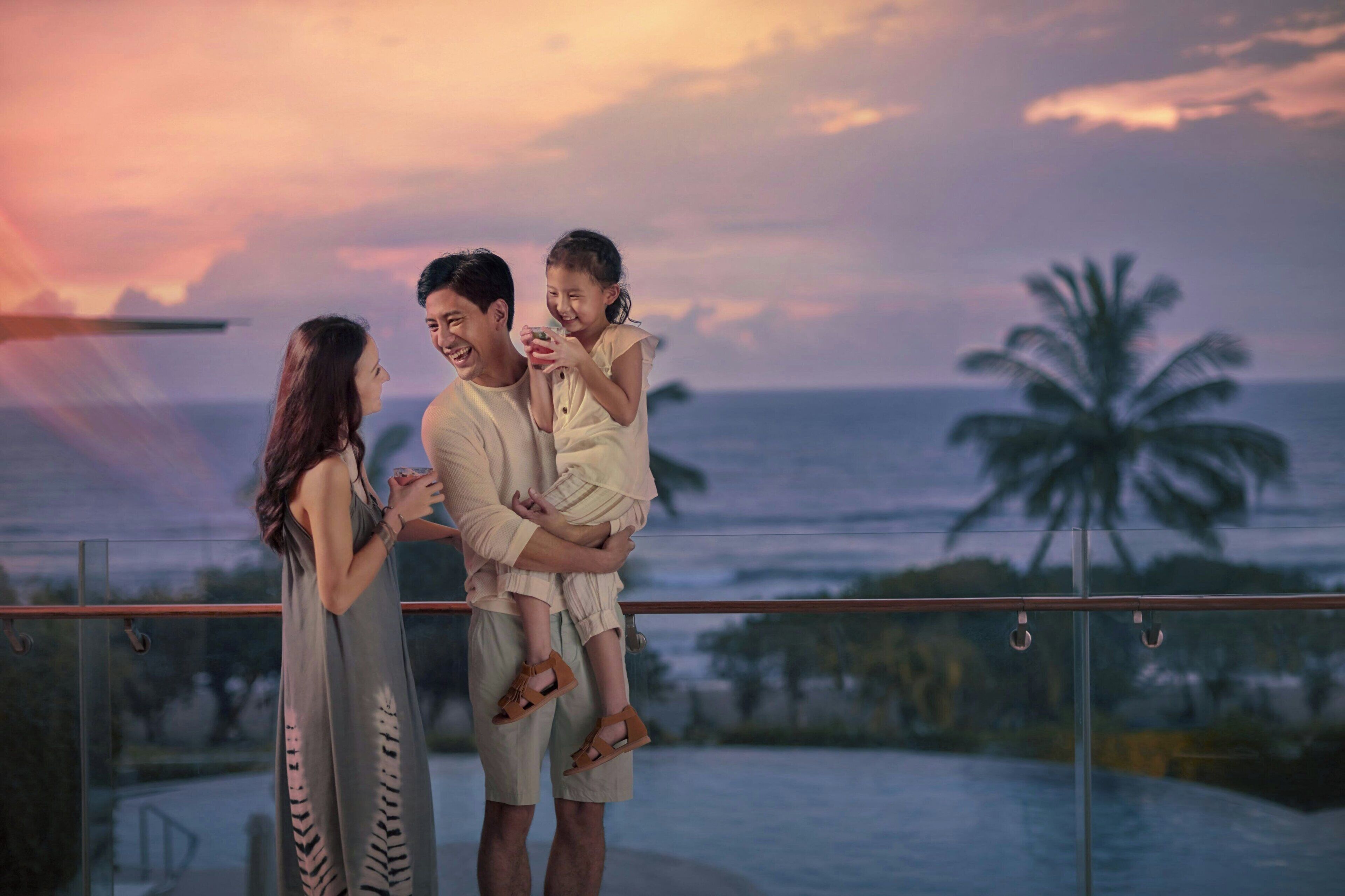 A couple holding a child on a balcony overlooking a body of water with palm trees and a sunset.