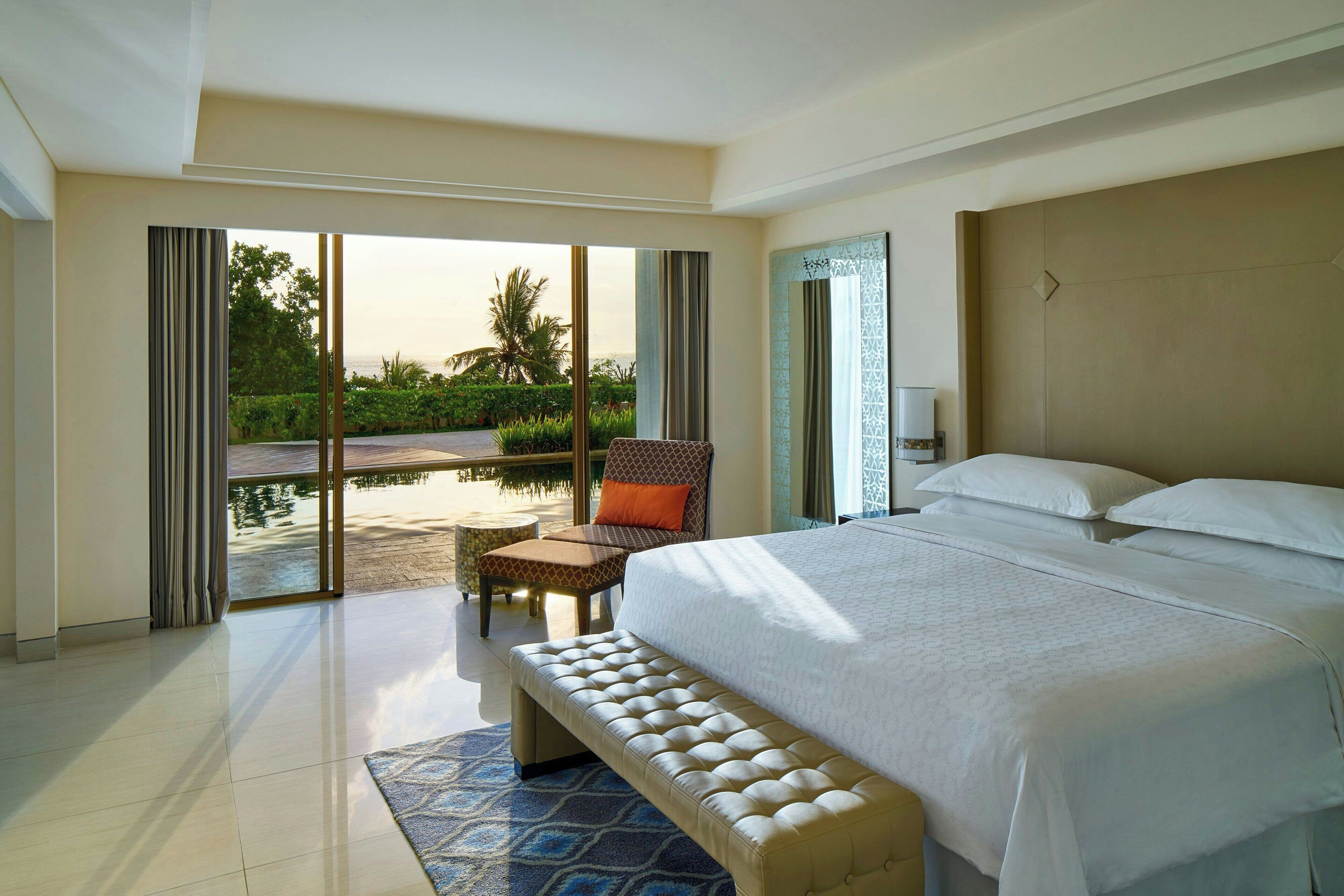 A hotel room with a large bed, a bench, a chair, and a view of a pool and greenery.