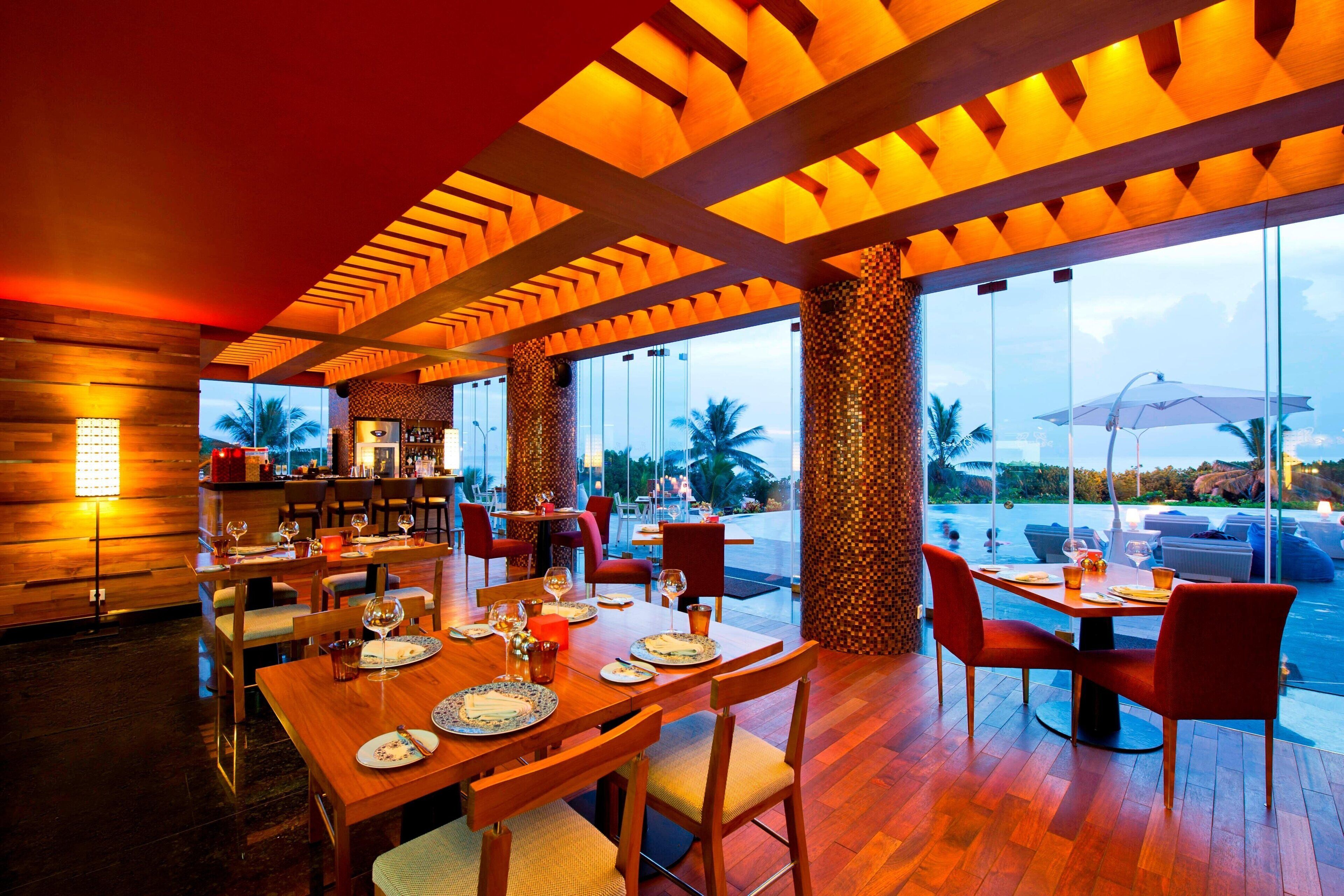 A restaurant with wooden tables and chairs, large windows, and a view of palm trees.
