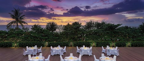 An outdoor dining area with round tables and white chairs, set against a backdrop of a sunset over the ocean.