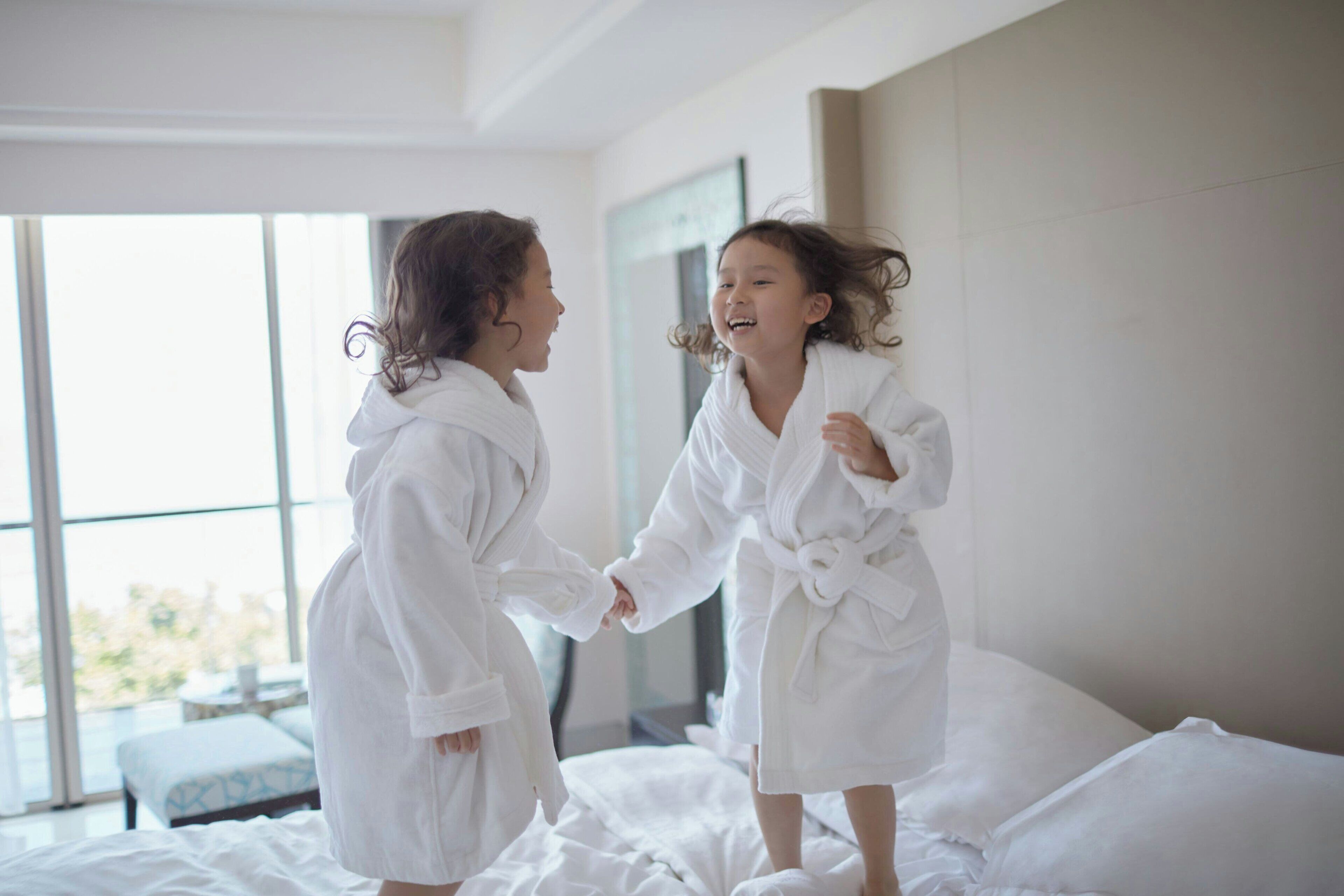 Dos personas con túnicas blancas sobre una cama en una habitación con ventanas amplias y vistas a la vegetación.