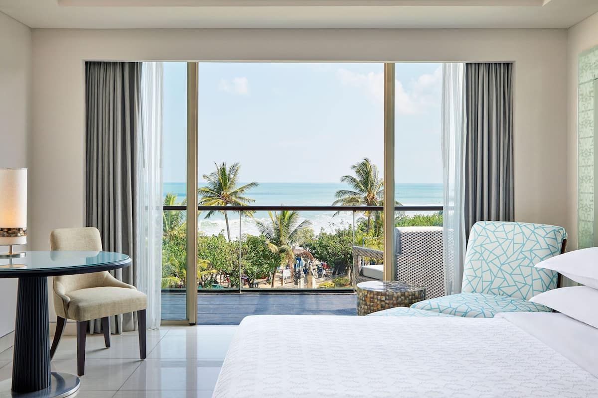 A hotel room with a large bed, a desk, and a chair. It has a balcony with a view of the beach and palm trees.