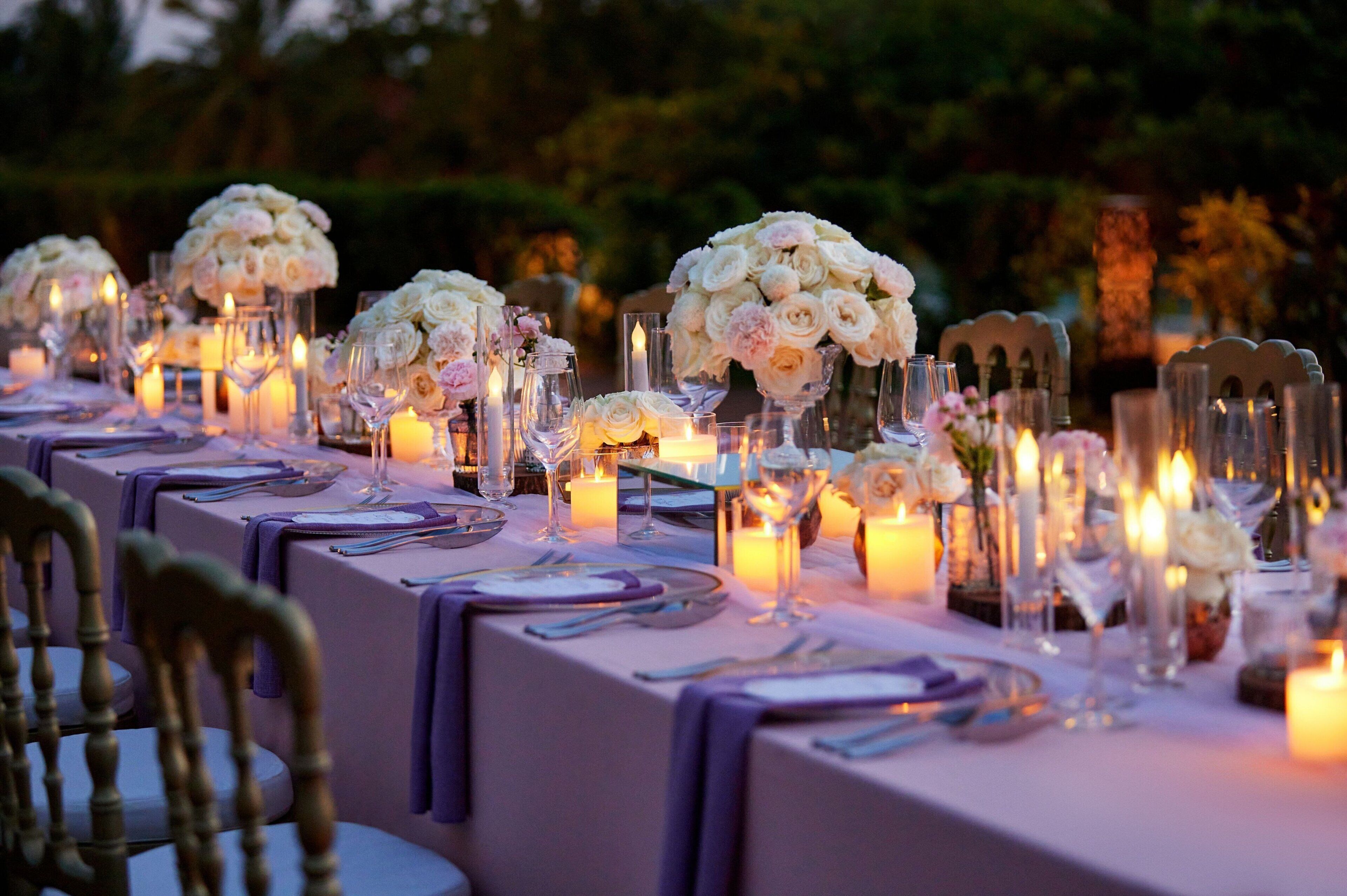 Una larga mesa dispuesta para un evento formal, con velas, copas y centros de mesa florales.