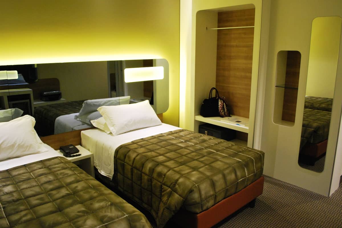 A hotel room with two beds, a desk with a lamp, a mirror, a wardrobe, and a handbag on the nightstand.