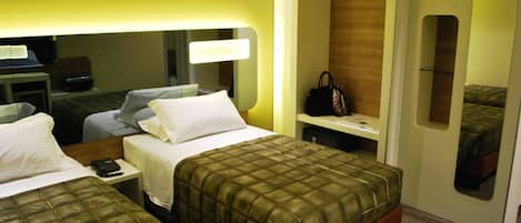Habitación de hotel con dos camas, un escritorio con lámpara, un espejo, un armario y un bolso sobre la mesita de noche.