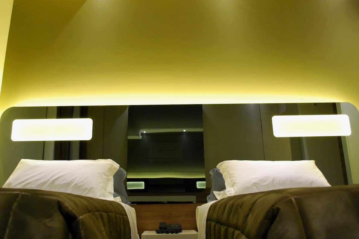 A hotel room with two beds, each with a pillow and a blanket. There are bedside tables with lamps and a small table between the beds.