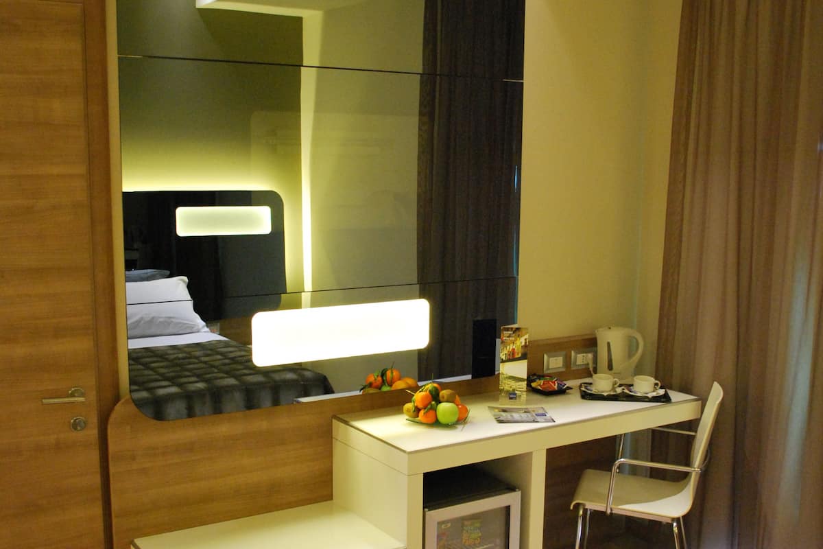 A compact hotel room with a bed, a desk, a chair, a mirror, and a small kitchenette area.