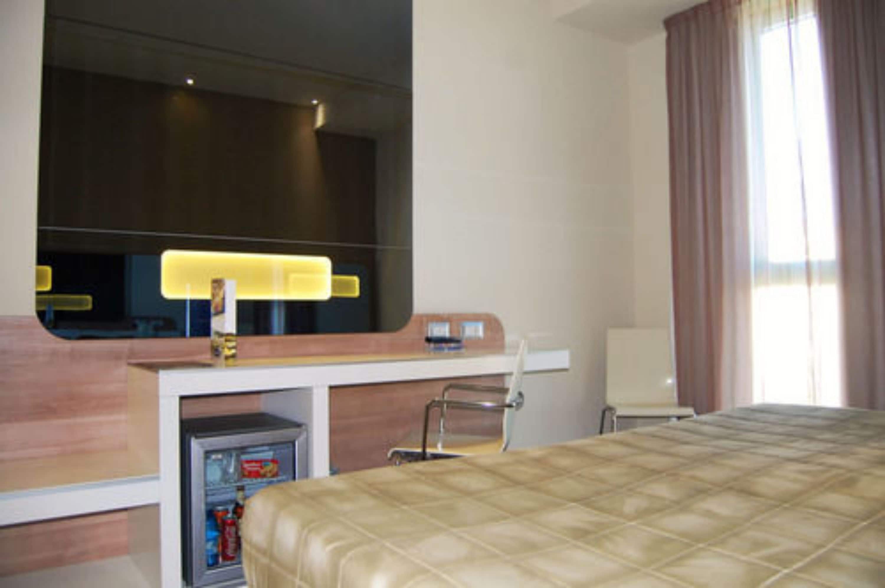 A hotel room with a bed, a desk with a computer, a chair, a minibar, and a large mirror.