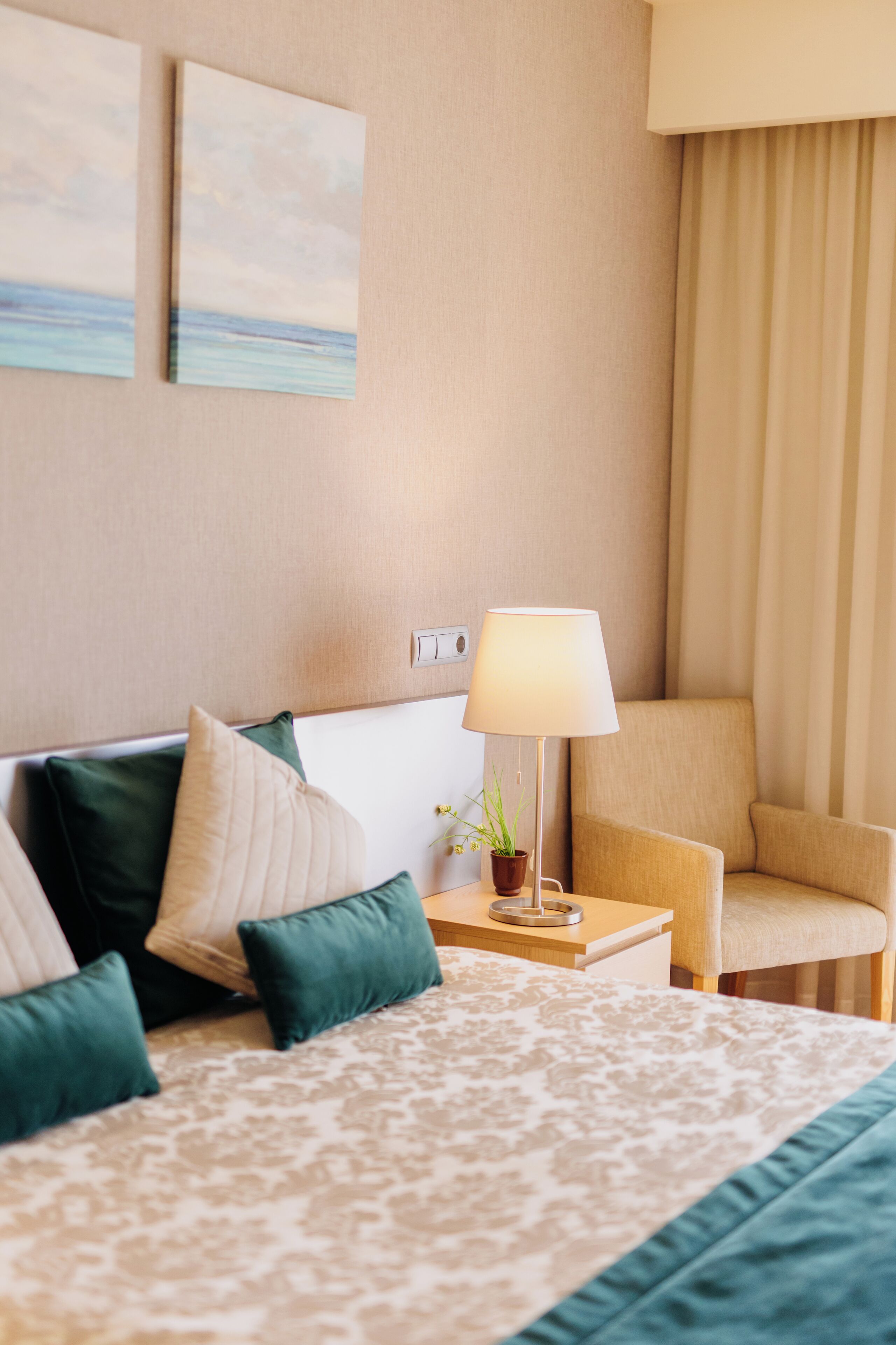 Quarto de hotel com cama, uma cadeira, um abajur e duas pinturas na parede.