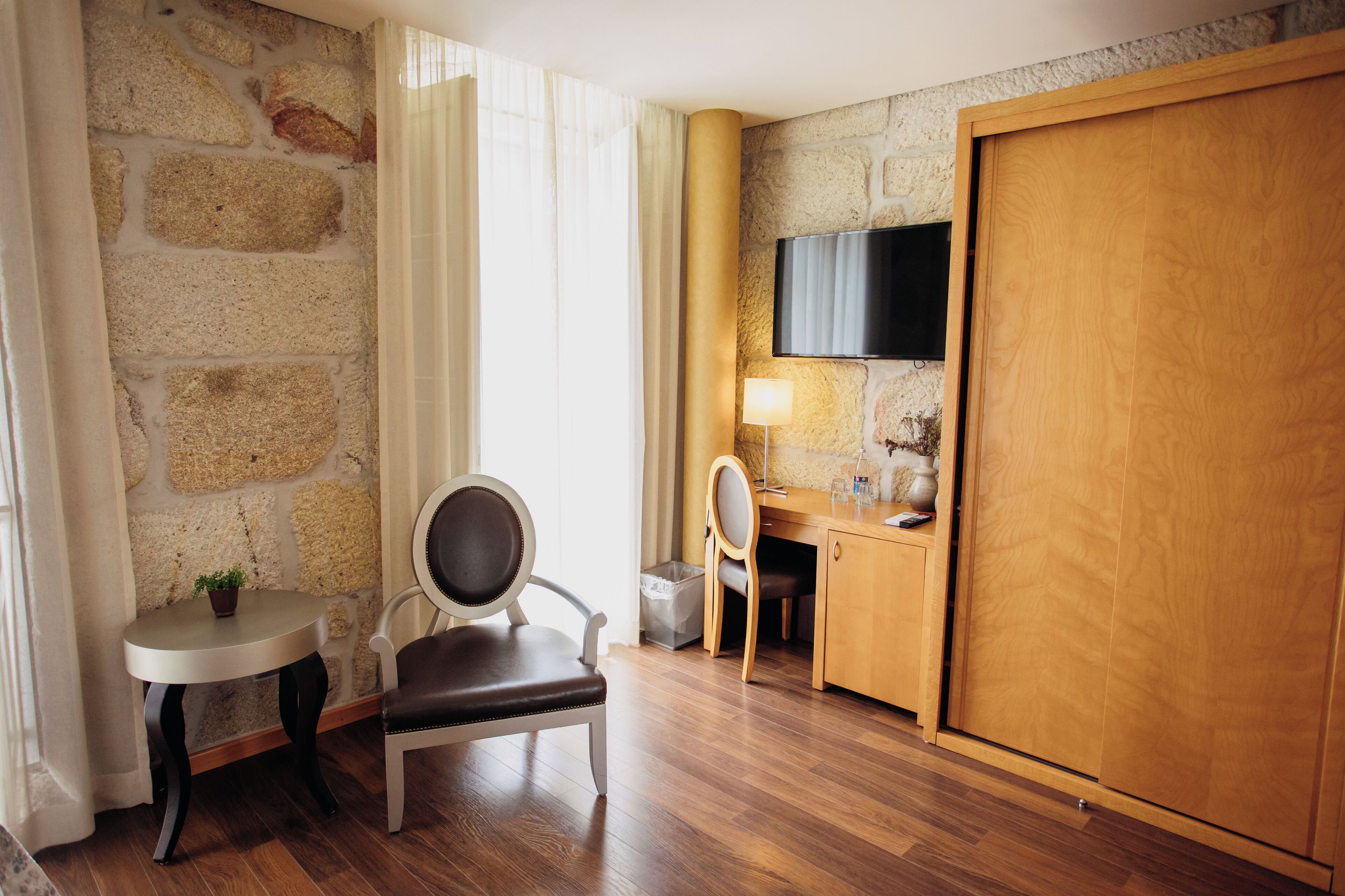 Een hotelkamer met een stenen muur, een houten bureau, een stoel, een aan de muur bevestigde televisie en een houten garderobe.