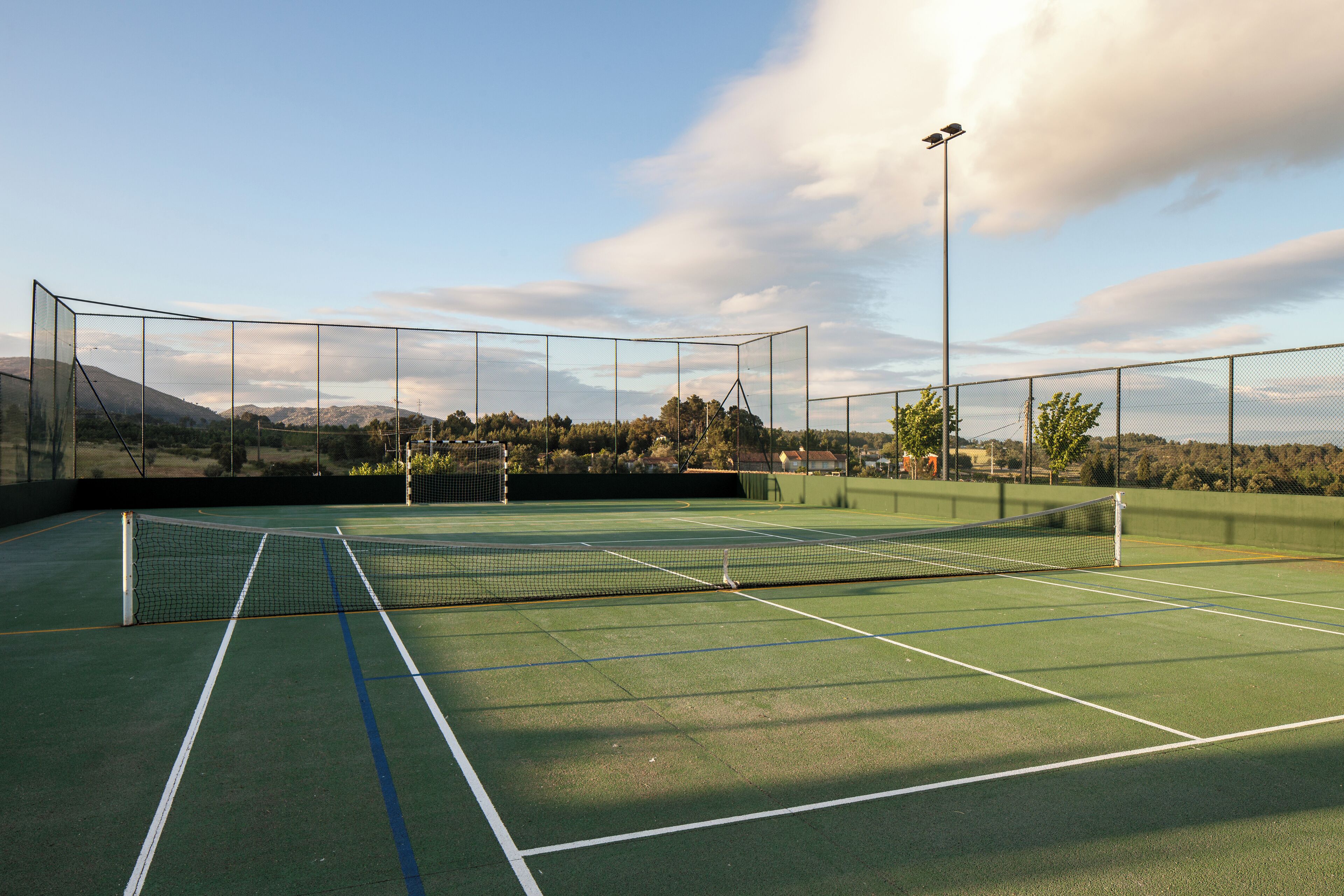 An outdoor tennis court with a net, surrounded by a fence, and a clear sky with clouds.