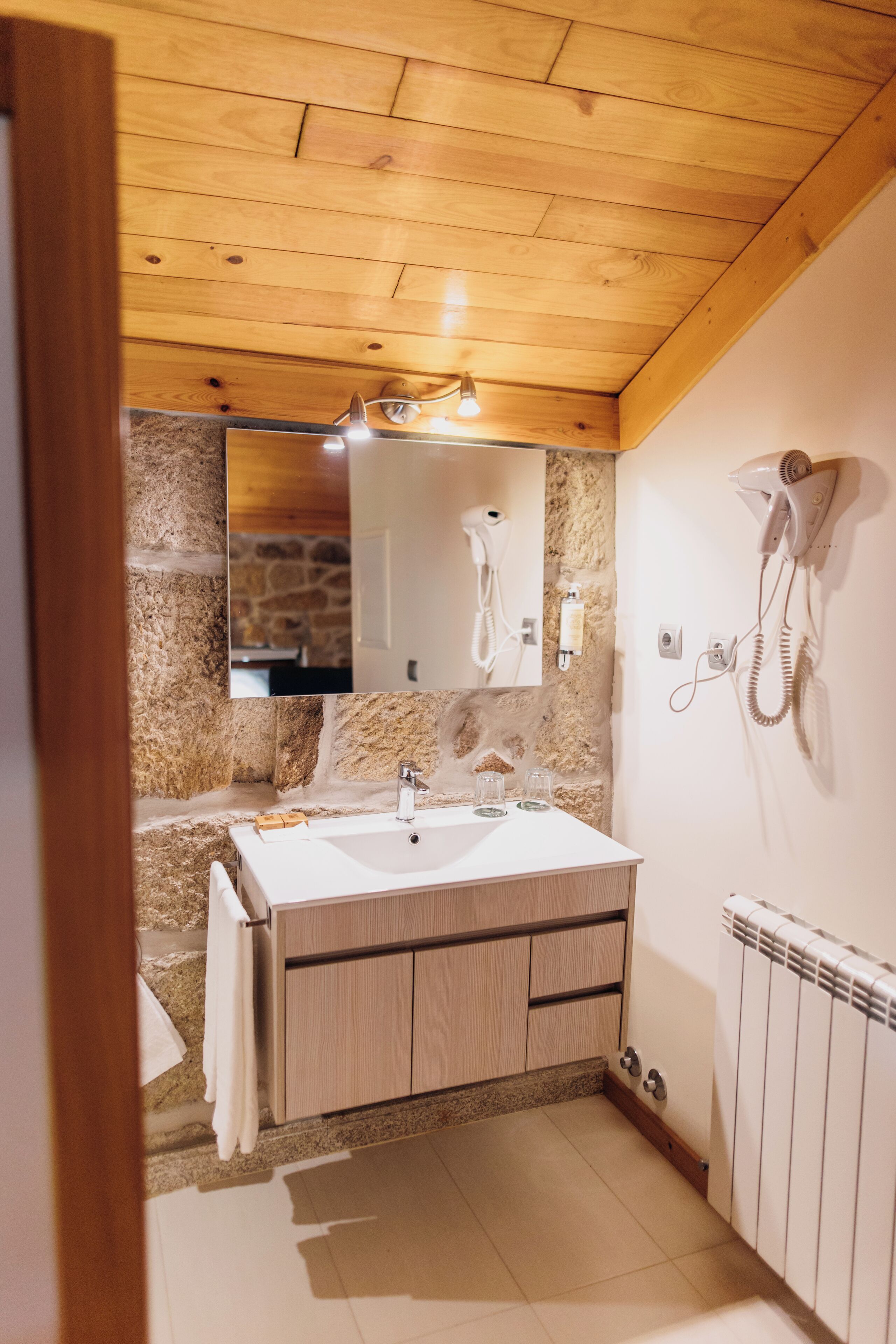 A bathroom with a wooden ceiling, stone wall, two sinks, a mirror, a hairdryer, and a towel rack.