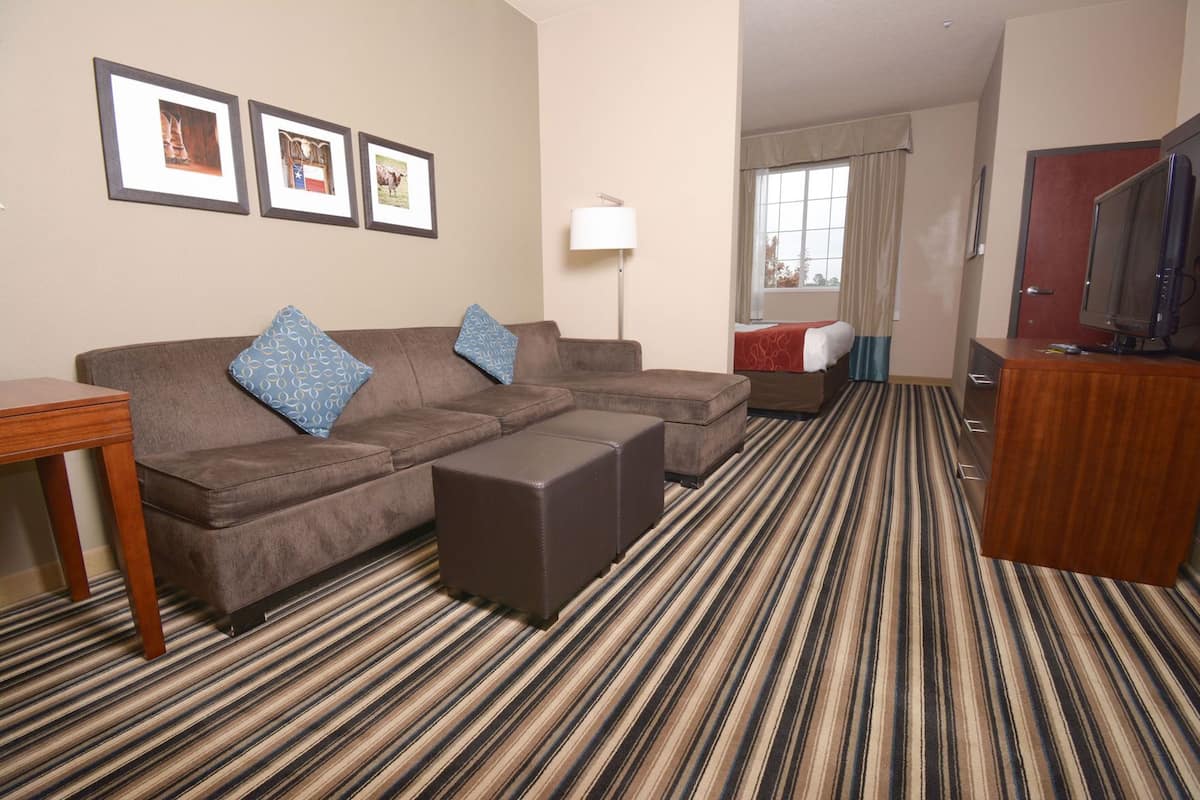 A hotel room with a sectional sofa, ottoman, a TV, a lamp, and a window with curtains.