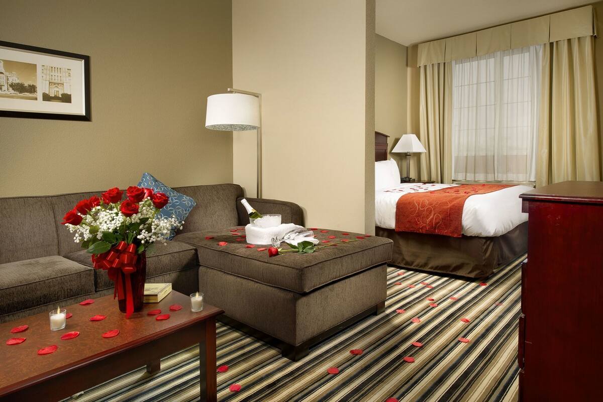 A hotel room with a sofa, a small table with a vase of flowers, a bed with a red blanket, and a large window with curtains.