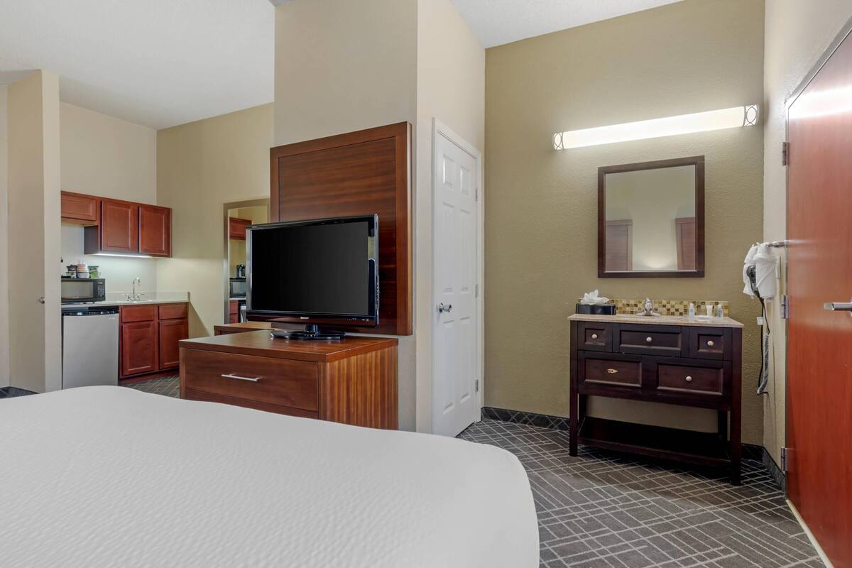 A hotel room with a bed, a TV, a wooden dresser, a mirror, a small kitchenette, and a bathroom.