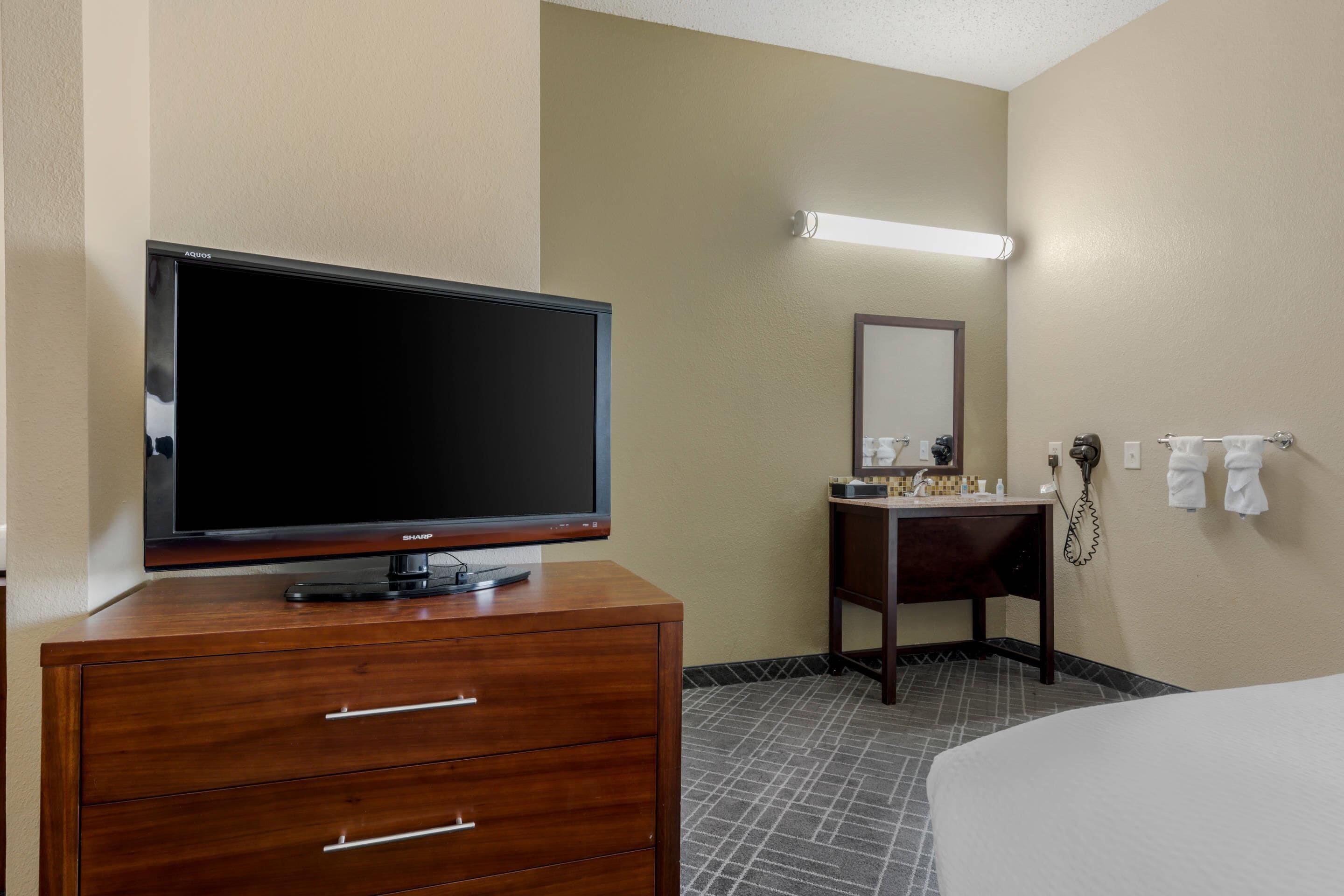 A hotel room with a flat-screen TV on a wooden dresser, a small vanity with a mirror, and a bed with a white comforter.