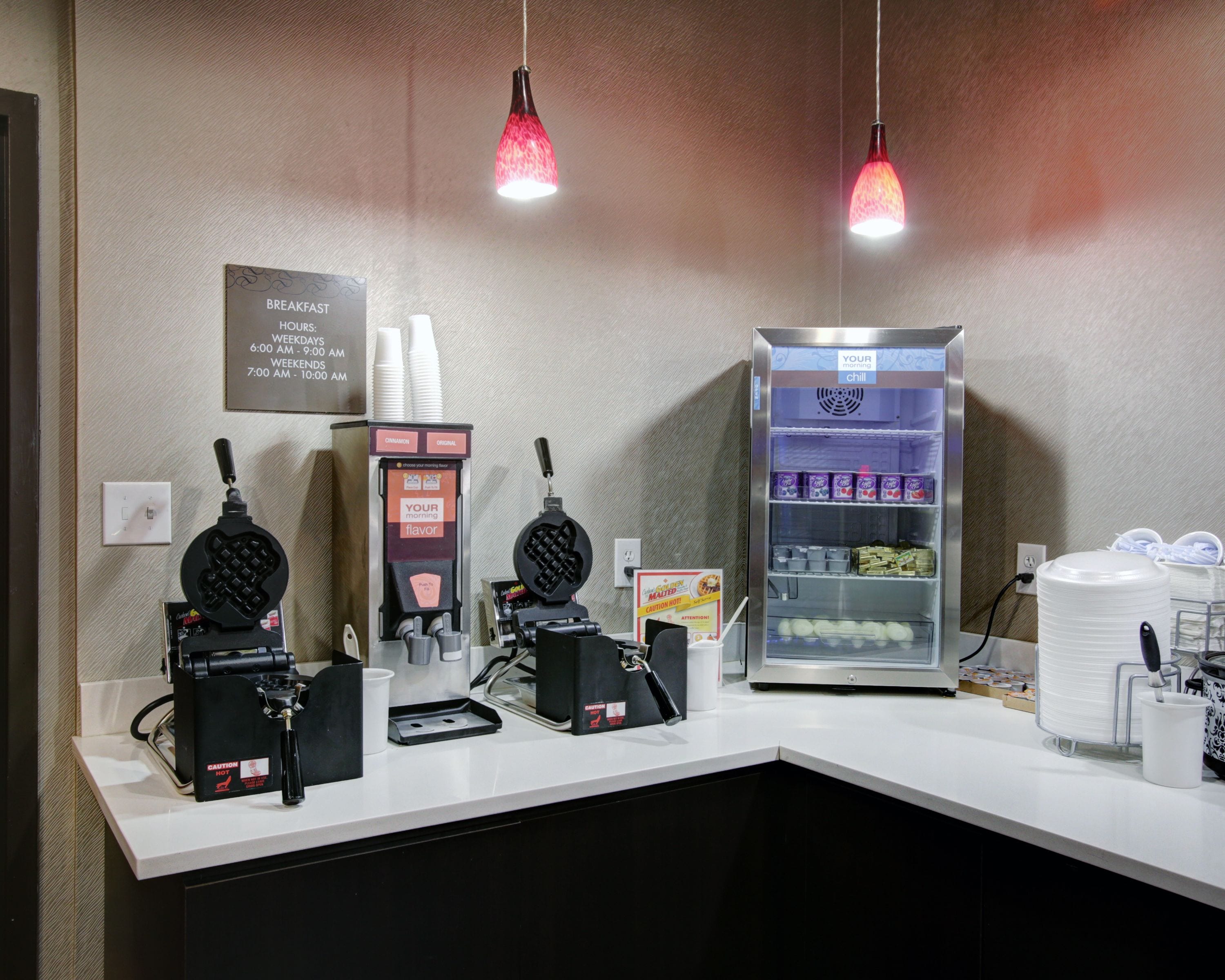 A breakfast station with waffle makers, a coffee machine, and a refrigerated drink dispenser.