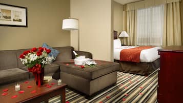 A hotel room with a sofa, a small table with a vase of flowers, a bed with a red blanket, and a large window with curtains.