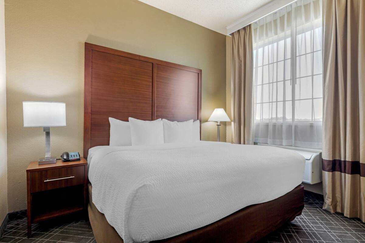 A hotel room with a large bed, two bedside tables with lamps, a window with curtains, and a wall-mounted heater.