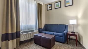 A hotel room with a blue sofa, a small table with a lamp, a window with curtains, and a radiator.