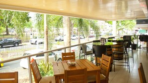 Un espace extérieur aménagé, abrité et doté de tables et de chaises en bois, avec de grandes fenêtres donnant sur une rue animée, où l’on peut voir des voitures et des palmiers.