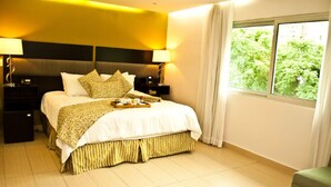 Une chambre d’hôtel avec un grand lit, deux tables de chevet avec des lampes, une fenêtre donnant sur un paysage verdoyant et un fauteuil vert.