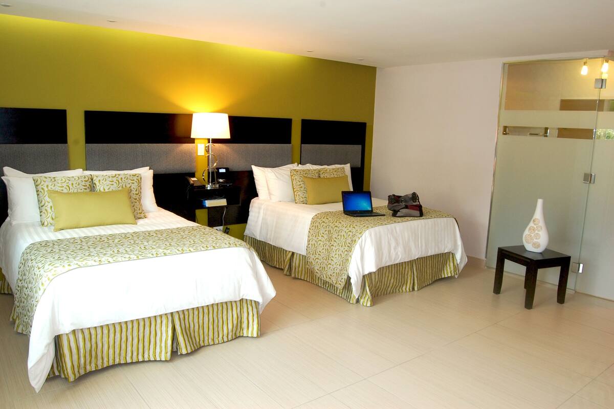 A hotel room with two beds, a nightstand with a lamp, a small table with a vase, and a wardrobe.