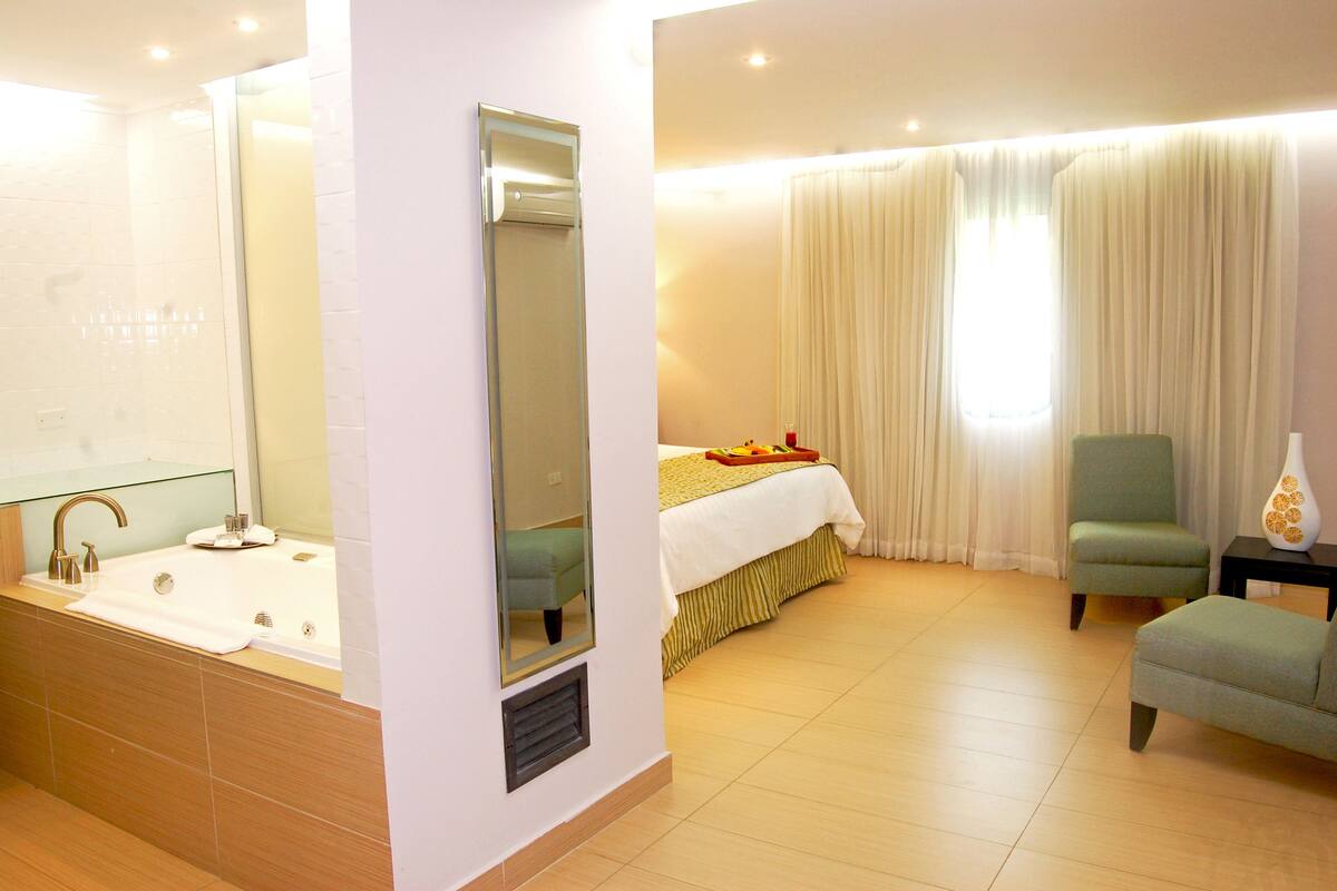 A hotel room with a large bathroom, a bed with a white bedspread, a green armchair, and a small table with a vase.