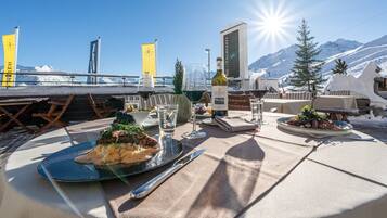 A dining table set with plates of food, glasses of wine, and a bottle of wine, with a mountain backdrop and flags.