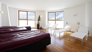 Habitación de hotel con una cama grande, dos sillones blancos, una planta en maceta y ventanas amplias con vistas a la montaña.