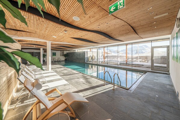 A modern indoor pool area with wooden ceiling, glass walls, and lounge chairs. - Hotel Hochsölden (Soelden)