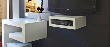 Um espaço de estar moderno, com uma TV de tela plana presa a uma parede escura, um móvel branco embaixo e uma pequena lixeira preta.