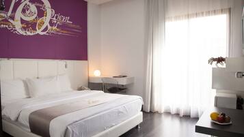 Una habitación de hotel moderna con una cama grande, un escritorio con lámpara, un frutero y una pared morada con texto decorativo.