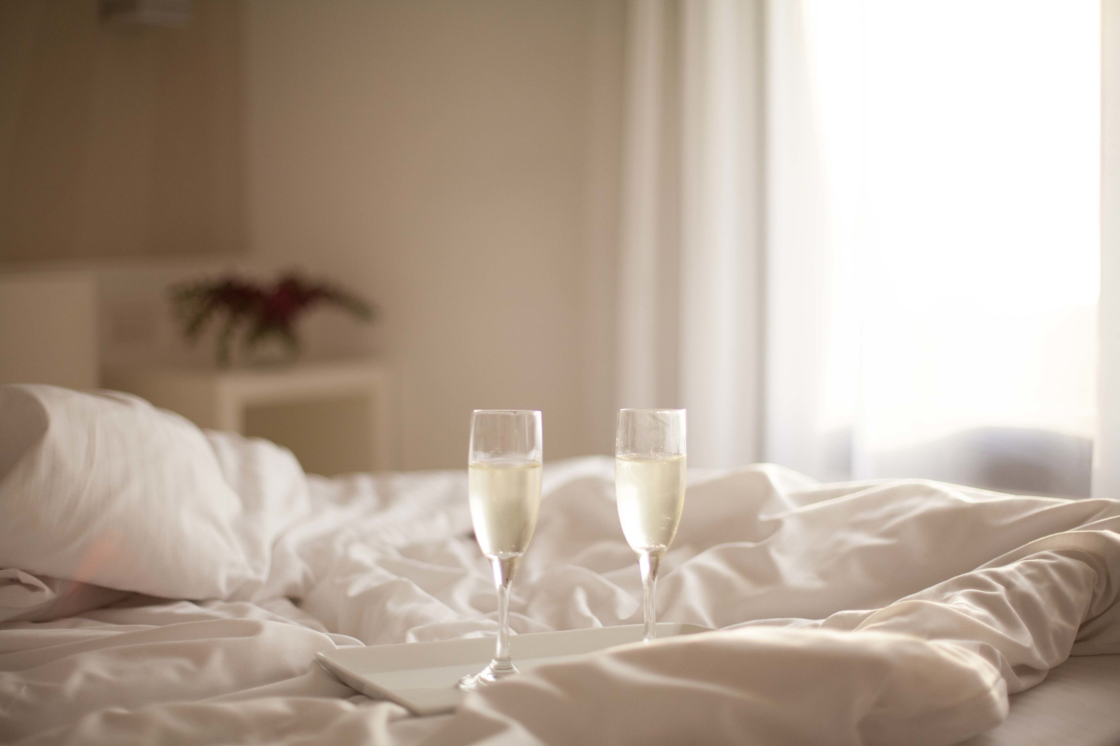 A bed with two glasses of champagne, a white pillow, and a bedside table with a plant.