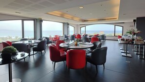 A modern dining area with round tables and chairs, large windows offering a view of the sea, and a ceiling with recessed lighting.
