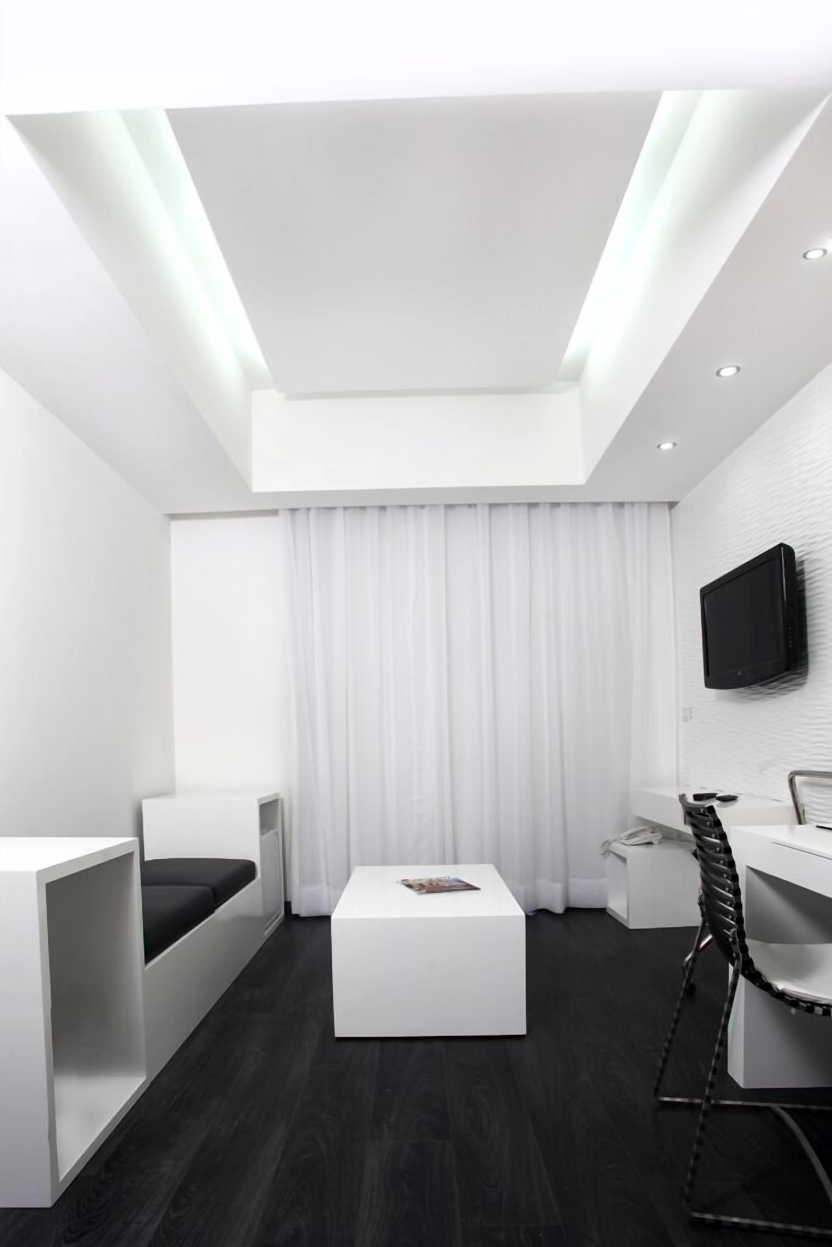 Quarto de hotel moderno com cama branca, escrivaninha branca, TV de tela plana na parede e pequena mesa de apoio.