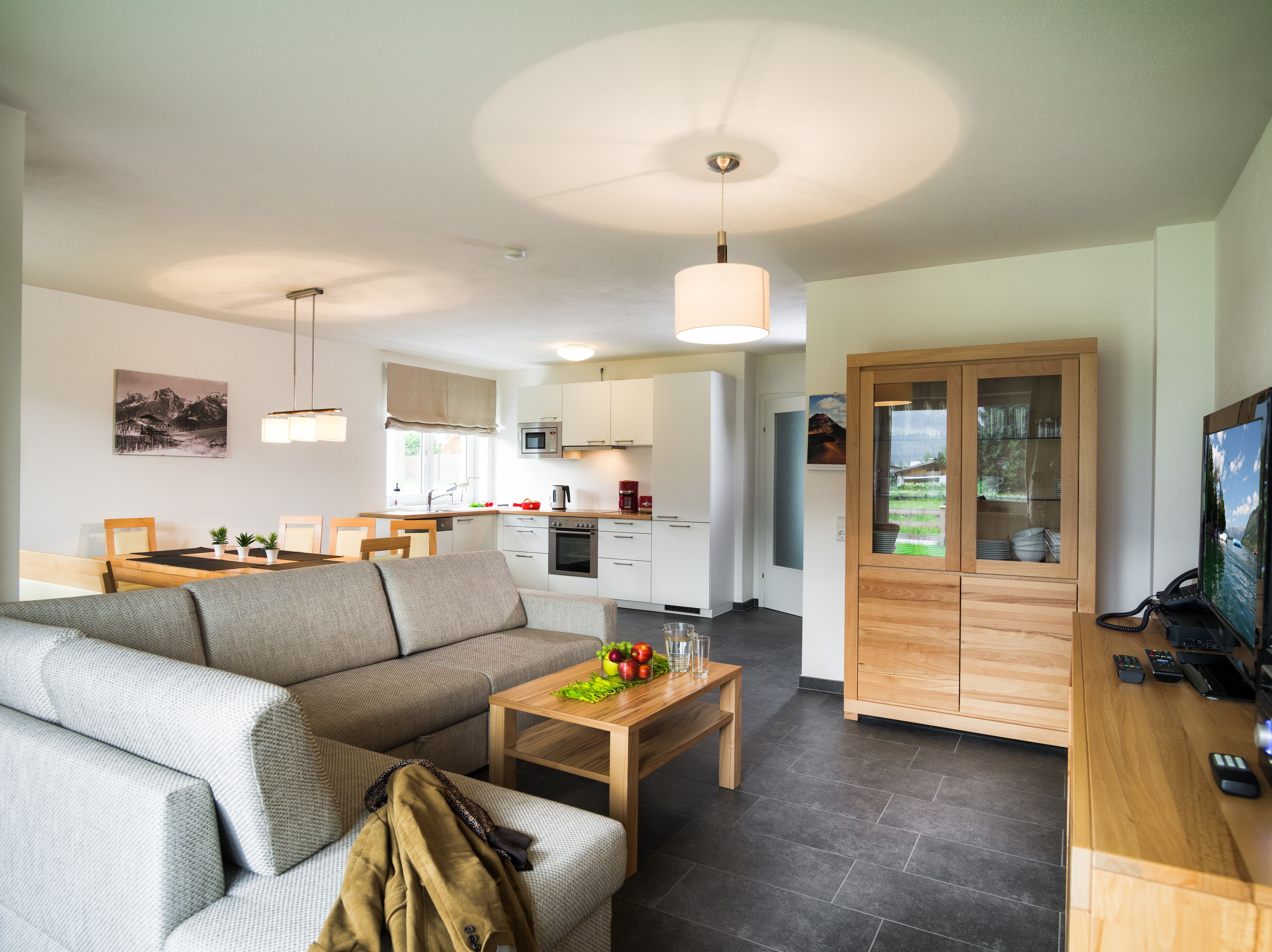 A modern living room with a sofa, coffee table, and a TV. There is a kitchen area with cabinets and a microwave.