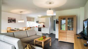 A modern living room with a sofa, coffee table, and a TV. There is a kitchen area with cabinets and a microwave.