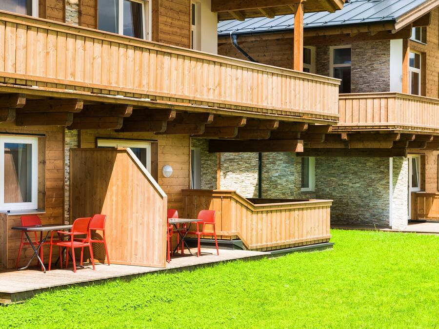 A multi-story wooden building with a balcony, outdoor seating area with red chairs, and a stone facade.