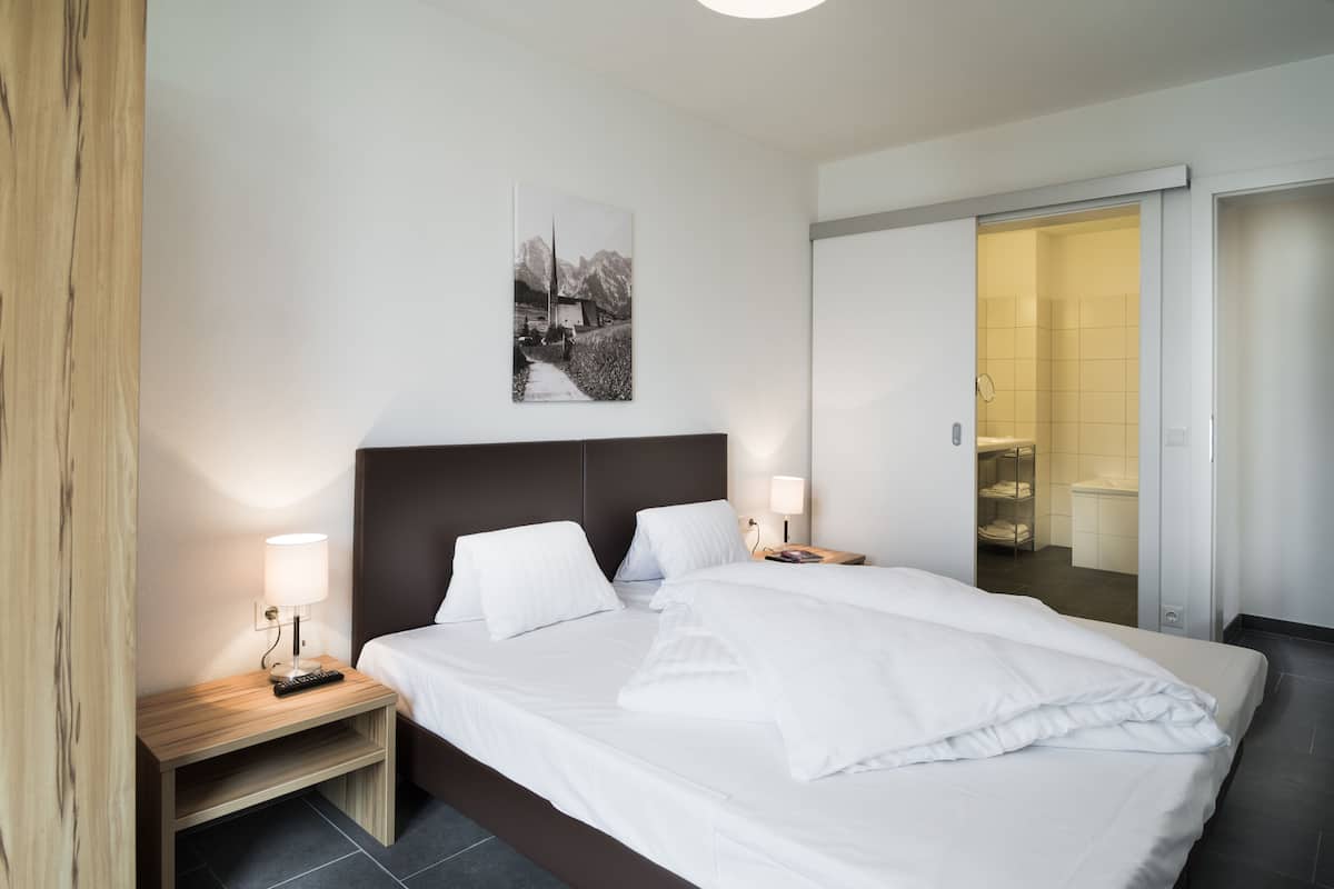 Une chambre d’hôtel moderne avec un grand lit, des tables de chevet, un tableau accroché au mur et une porte coulissante donnant accès à la salle de bain.
