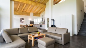 Een ruime woonkamer met houten plafond, een grote hoekbank, een houten salontafel, en een keuken op de achtergrond.