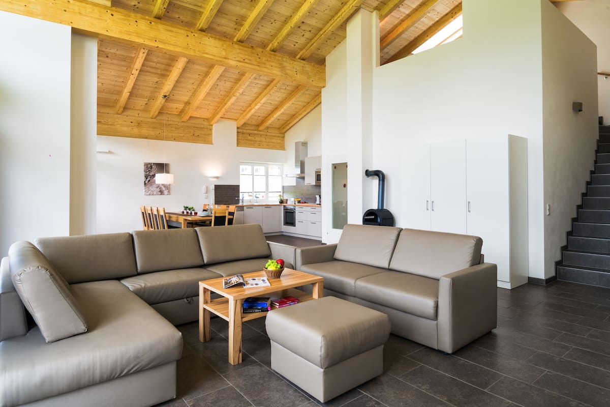 Un vaste salon doté d’un plafond en bois, d’un grand canapé en forme de L, d’une table basse en bois et d’une cuisine à l’arrière-plan.